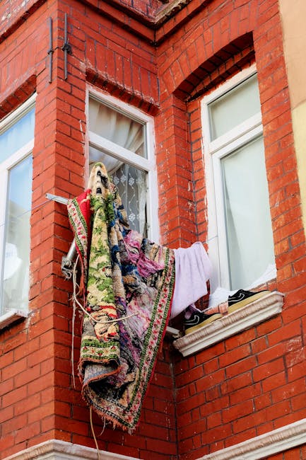 Exterior view of a red brick residential building with two white-framed sash windows. A small clothes drying rack is positioned outside the window, holding various textiles including a patterned carpe